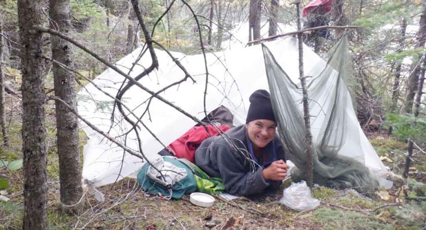 a student smiles from a tarp shelter in a wooded area beside a body of water 